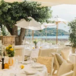 Set tables beneath a large tree and parasols with the bay behind at Loulou Restaurant Saint-Tropez