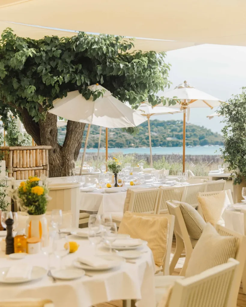 Set tables beneath a large tree and parasols with the bay behind at Loulou Restaurant Saint-Tropez
