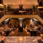 Booth dining beneath a mezzanine with a bronze staircase and pendant lights at Manko Paris