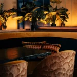 Curved banquette with printed cushions and carved chairs below lit foliage at Manko Paris