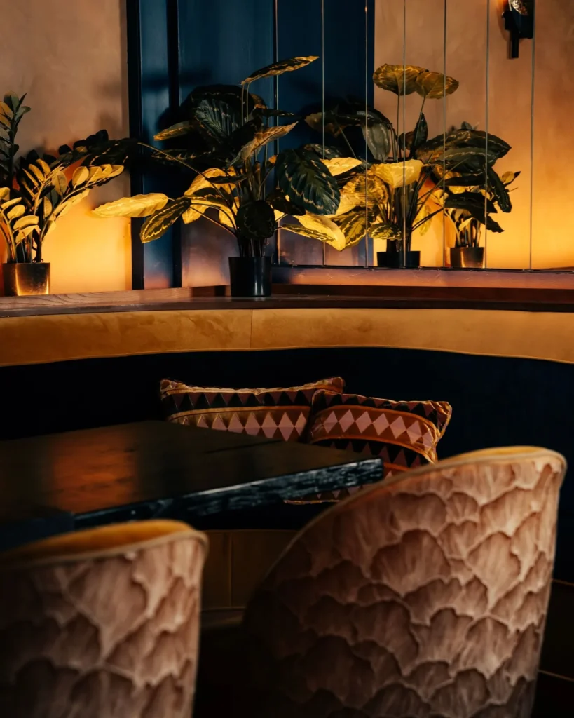 Curved banquette with printed cushions and carved chairs below lit foliage at Manko Paris
