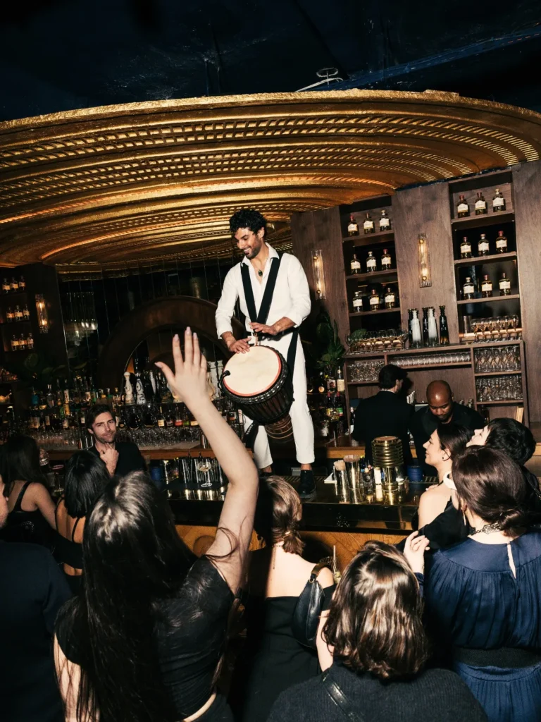 Performer in white playing a djembe atop the bar before a crowd under the gold ceiling at Manko Paris