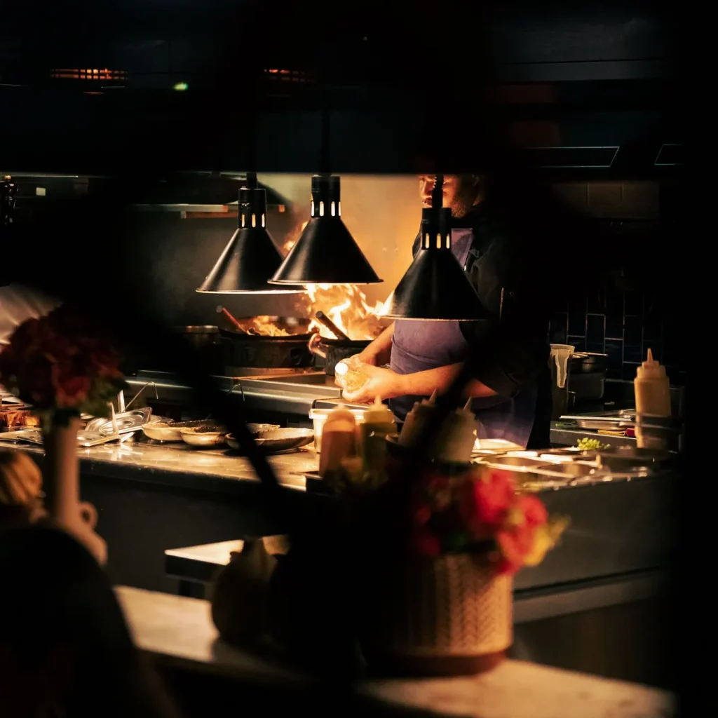Chef cooking over flames beneath black heat lamps in the open kitchen at Manko Paris