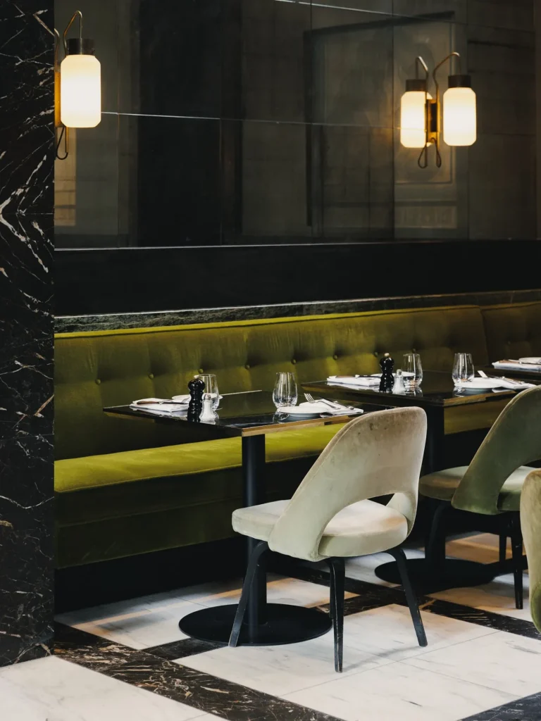 Green banquette and marble table below glass wall sconces on a checkerboard floor at Monsieur Bleu Paris