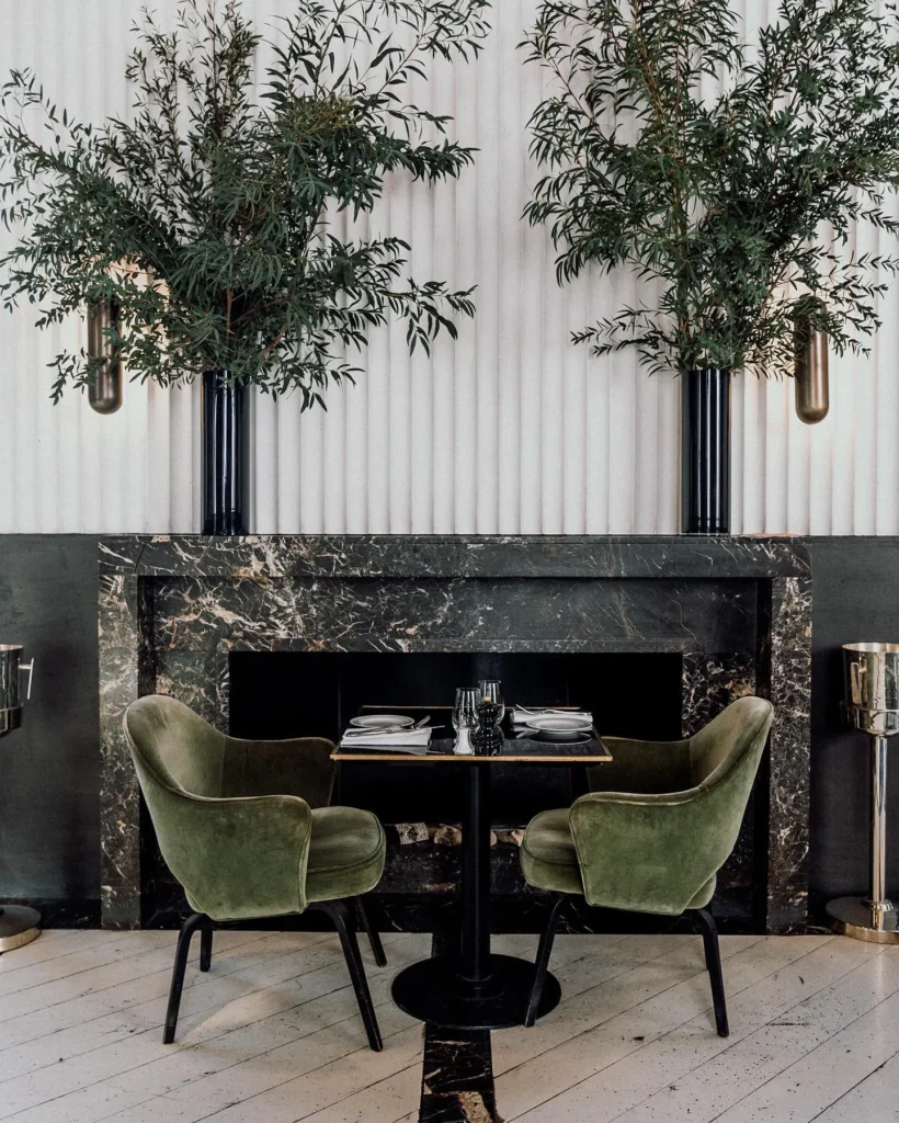 Green velvet chairs at a table set within a marble fireplace below tall olive branches at Monsieur Bleu Paris