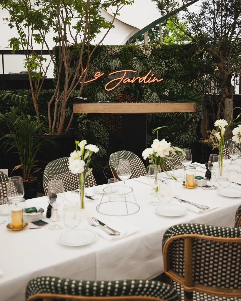Long event table set before a Le Jardin neon sign and DJ booth in a greenery-filled space at Monsieur Bleu Paris