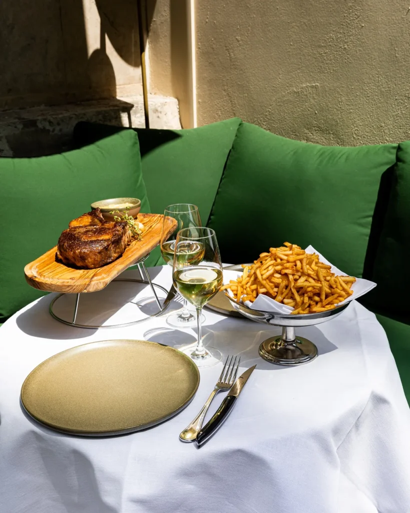 Grilled rib steak on a wooden board with a silver dish of fries on a terrace table at Monsieur Bleu Paris