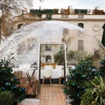 Transparent geodesic dining dome with a fur-draped table flanked by decorated fir trees by day at Monsieur Bleu Paris