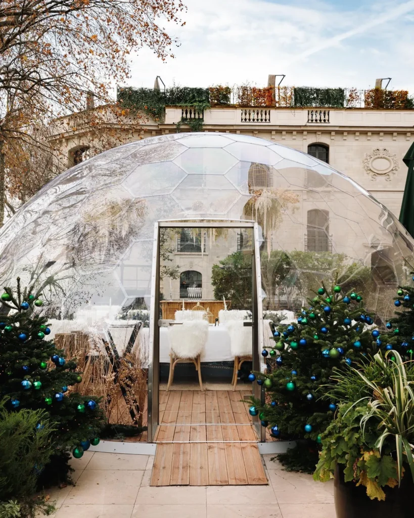 Transparent geodesic dining dome with a fur-draped table flanked by decorated fir trees by day at Monsieur Bleu Paris