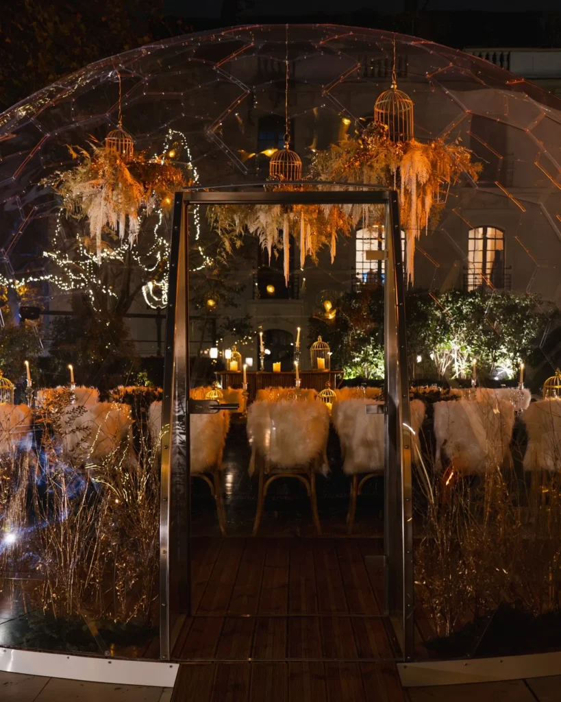 Illuminated winter dining dome with hanging birdcages and fur-draped chairs at night at Monsieur Bleu Paris