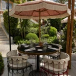 Round café table with skirted chairs under a pink fringed parasol in the garden at Monsieur Dior des Lices Saint-Tropez by Mauro Colagreco
