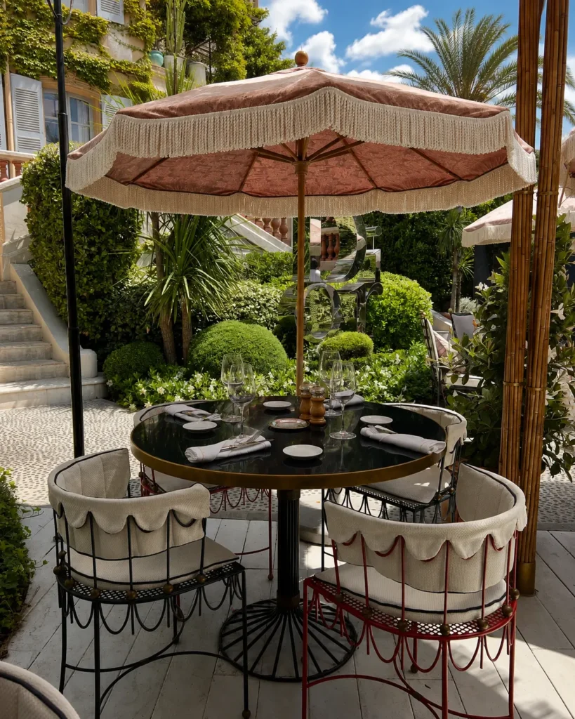 Round café table with skirted chairs under a pink fringed parasol in the garden at Monsieur Dior des Lices Saint-Tropez by Mauro Colagreco