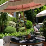 Café table set under a pink fringed parasol beside the mirrored Dior sculpture at Monsieur Dior des Lices Saint-Tropez by Mauro Colagreco