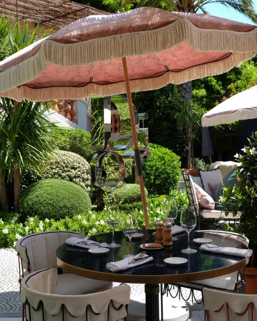 Café table set under a pink fringed parasol beside the mirrored Dior sculpture at Monsieur Dior des Lices Saint-Tropez by Mauro Colagreco