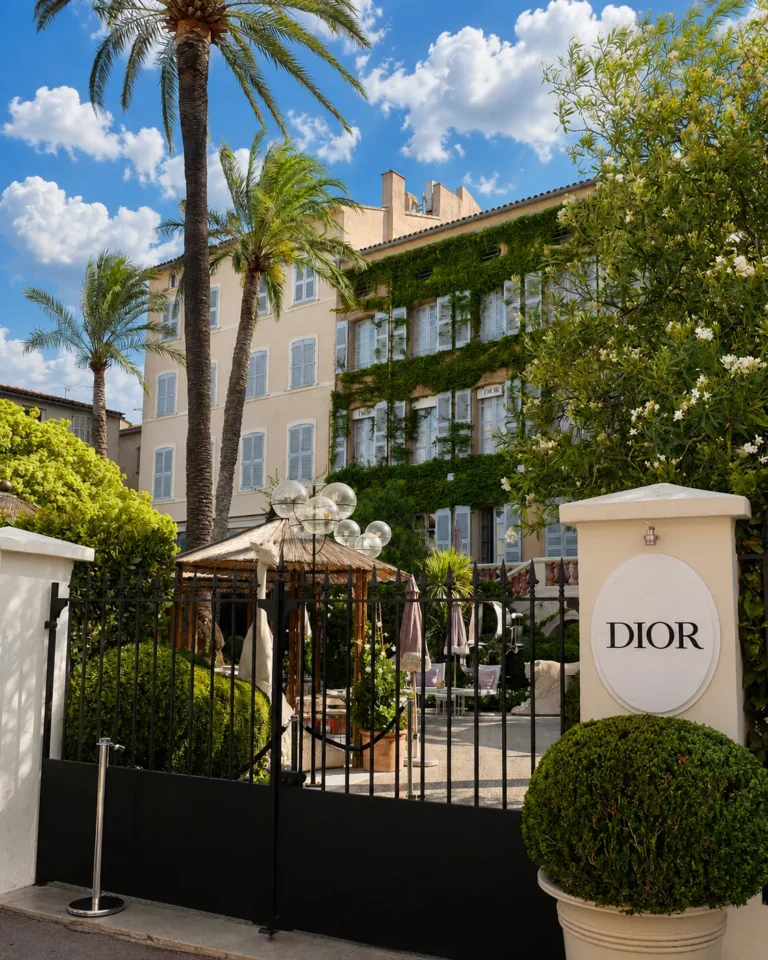 Iron gate with an oval DIOR sign and palm trees before the ivy-clad facade at Monsieur Dior des Lices Saint-Tropez by Mauro Colagreco