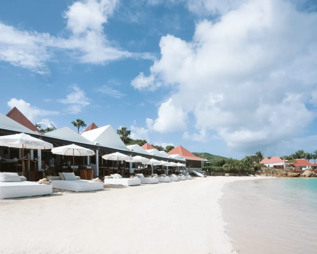 Row of white daybeds along the sand beside the red-roofed beach club and calm bay at Nikki Beach Saint Barth