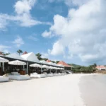 Row of white daybeds along the sand beside the red-roofed beach club and calm bay at Nikki Beach Saint Barth