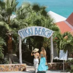 Two guests walking under the Nikki Beach arch sign framed by palms and turquoise sea at Nikki Beach Saint Barth