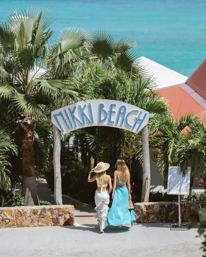 Two guests walking under the Nikki Beach arch sign framed by palms and turquoise sea at Nikki Beach Saint Barth