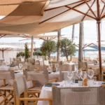 Restaurant tables under wooden parasols and olive trees with a sea view at Plage La Serena Ramatuelle Saint-Tropez