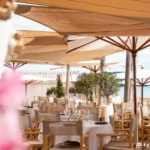 Restaurant tables beneath sail shades and parasols by the beach at Plage La Serena Ramatuelle Saint-Tropez