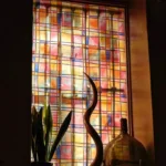 Backlit checkered stained-glass window with a kudu horn and demijohn on the sill at Restaurante El Cuerno Palma