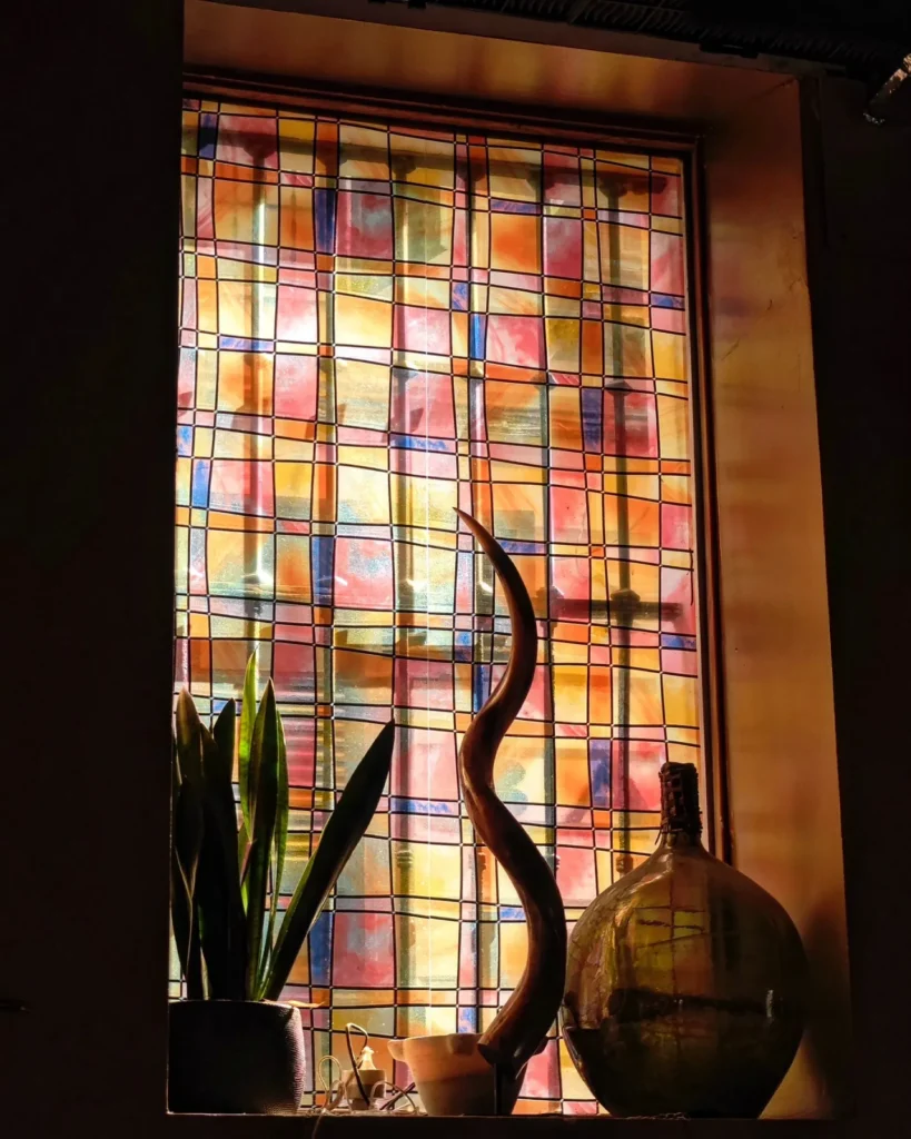 Backlit checkered stained-glass window with a kudu horn and demijohn on the sill at Restaurante El Cuerno Palma