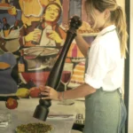 Server grinding pepper from an oversized mill over a dish beside a vintage poster at Rikito Palma