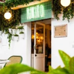 Green neon Rikito sign above the doorway framed by ivy and potted plants at Rikito Palma