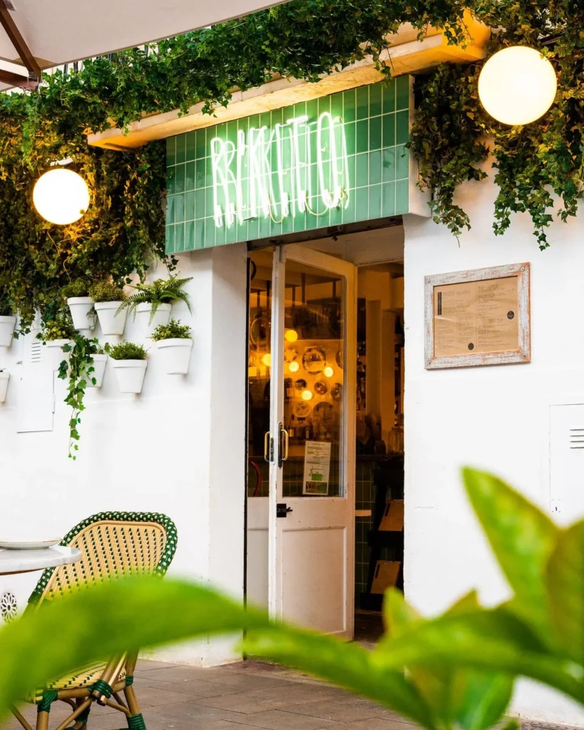 Green neon Rikito sign above the doorway framed by ivy and potted plants at Rikito Palma