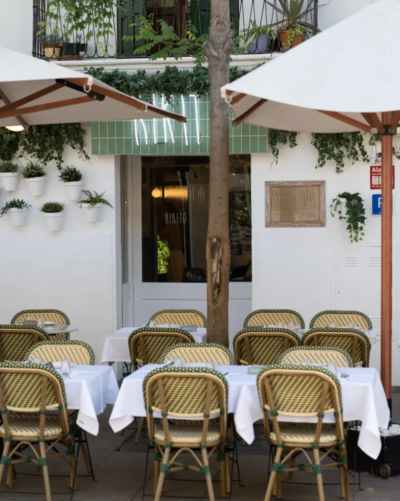 White-clothed terrace tables and bistro chairs around a tree beneath the Rikito neon sign at Rikito Palma