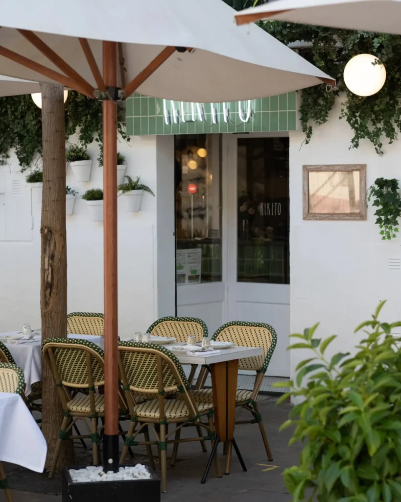 Terrace tables and green rattan bistro chairs under a parasol by the Rikito neon sign at Rikito Palma