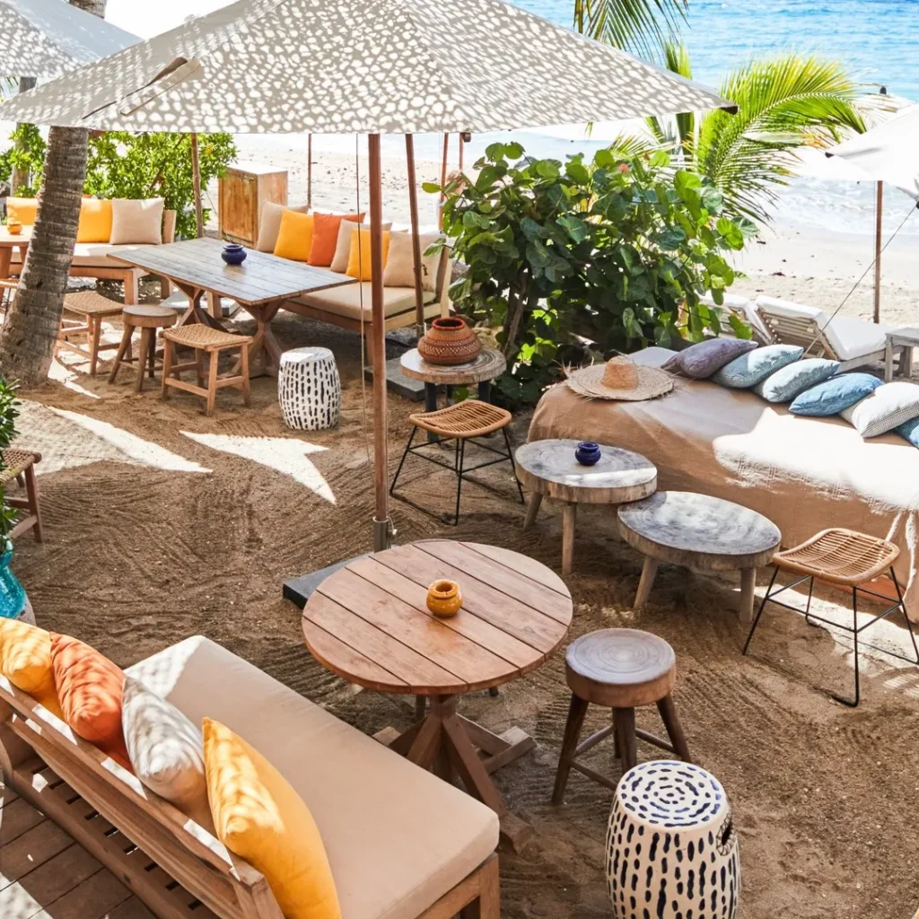 Cushioned sofas and low tables under a spotted umbrella beside the beach at Shellona Saint-Tropez