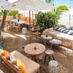 Cushioned sofas and low tables under a spotted umbrella beside the beach at Shellona Saint-Tropez