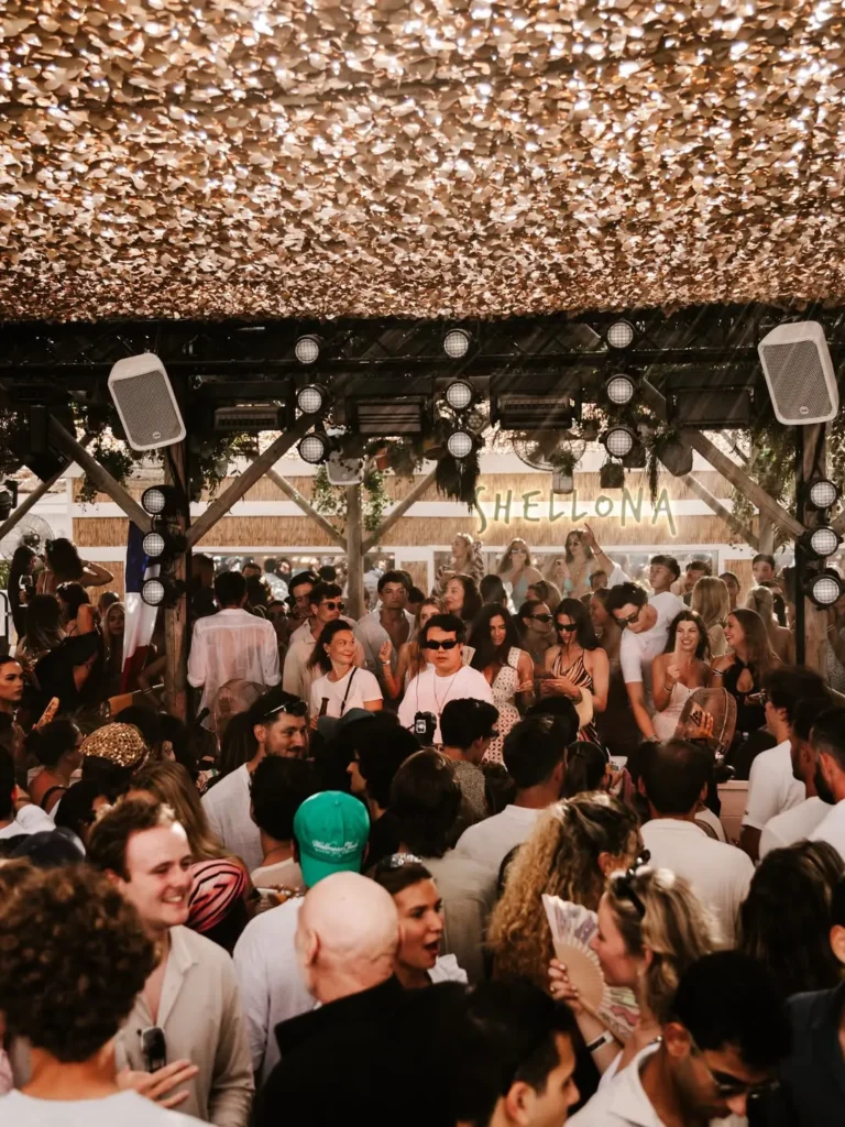 Packed crowd dancing beneath the lit Shellona sign and stage lights at Shellona Saint-Tropez