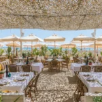 Beach restaurant tables under a woven canopy facing white parasols and the sea at Shellona Saint-Tropez