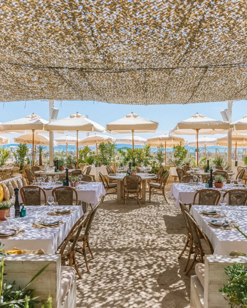 Beach restaurant tables under a woven canopy facing white parasols and the sea at Shellona Saint-Tropez