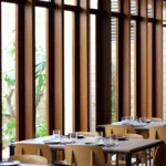 Dining tables and pale wood chairs along tall timber windows onto a garden at SPOT Palma De Mallorca