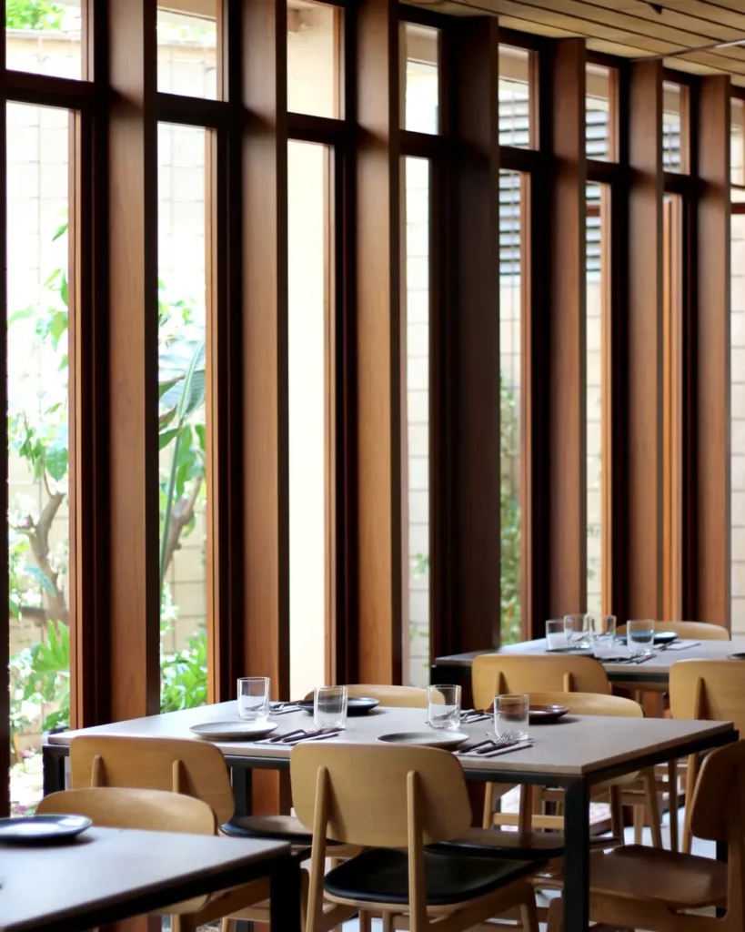 Dining tables and pale wood chairs along tall timber windows onto a garden at SPOT Palma De Mallorca