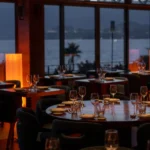 Set tables by full-height windows with a dusk sea view at Zuma Cannes