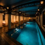 Indoor swimming pool at Hôtel Costes Paris, featuring turquoise-lit water, elegant curtained seating areas, dark wooden floors, decorative metal arches, and warm ambient lighting.”