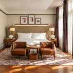 Warm contemporary hotel bedroom with a king-size bed, wood flooring, a comfortable lounge chair and sunlight streaming through tall windows