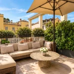 Sunny rooftop terrace with cushioned outdoor seating, a large parasol and lush greenery beside a Mediterranean-style building