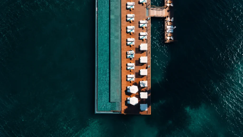 Aerial view of a long overwater resort pool surrounded by deep blue ocean and rows of lounge seating
