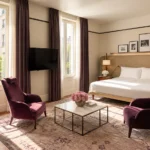 Elegant hotel bedroom with plum-colored armchairs, a king-size bed, framed artwork and warm neutral furnishings