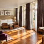 Bright luxury hotel suite with a comfortable seating area, elegant bedroom furnishings and large windows overlooking the city
