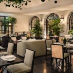 Refined dining lounge at The Carlton Milan with arched windows, leafy greenery, elegant tables, and upholstered chairs
