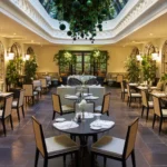 Elegant restaurant at The Carlton Milan arranged around a lush central atrium with a glass ceiling, greenery, and neatly set dining tables