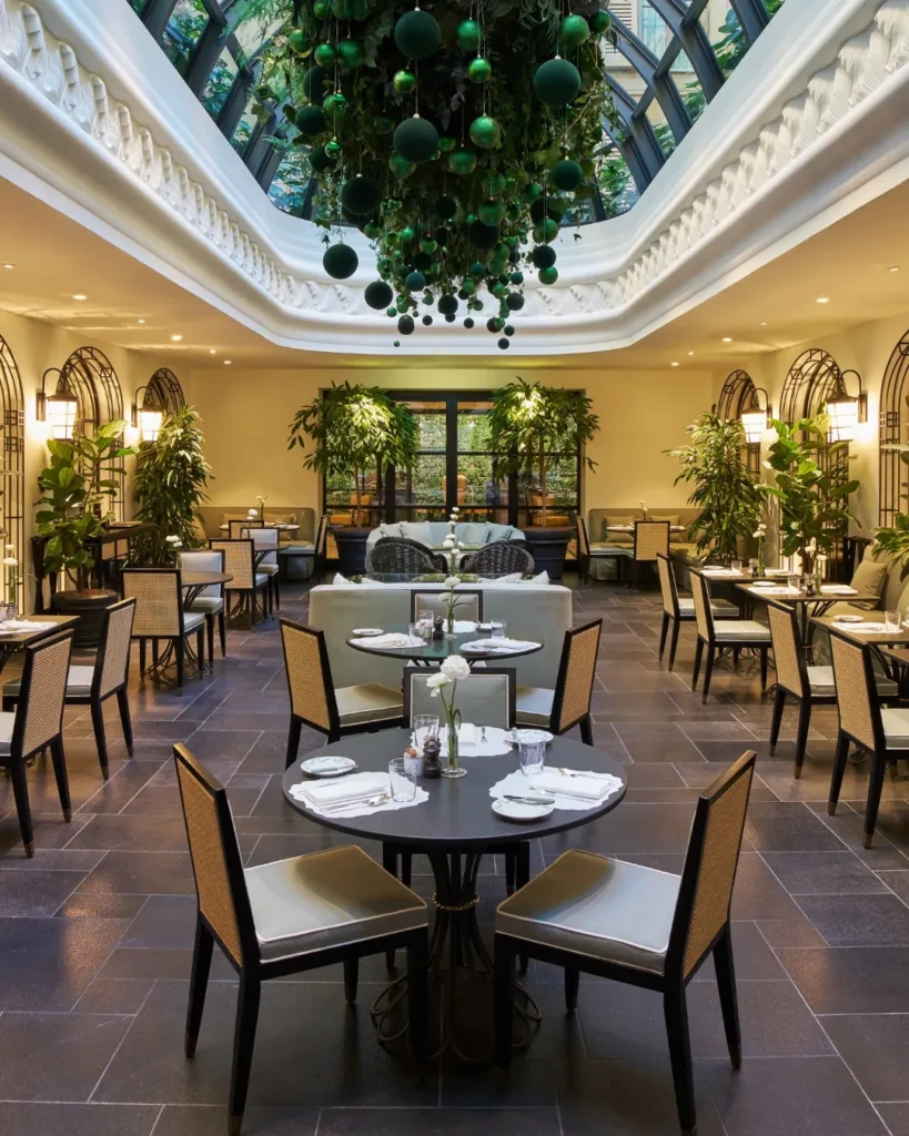 Elegant restaurant at The Carlton Milan arranged around a lush central atrium with a glass ceiling, greenery, and neatly set dining tables