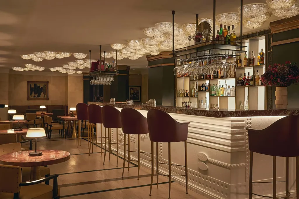 The Carlton Milan hotel bar featuring a backlit bottle display, elegant bar stools, sculptural ceiling lights, and contemporary furnishings